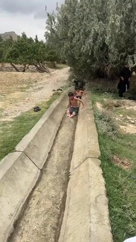 The video shows two young boys sliding down a concrete irrigation channel filled with water. The boy in the foreground is sitting on his bottom, sliding on his bare skin, while the boy behind him is in a more crouched position. Both boys appear to be wearing bathing suits or shorts. The channel is surrounded by dry, dusty ground and patches of green grass, with trees in the background. In the far background, there are some hills or mountains. The sky is overcast, suggesting a cloudy or possibly rainy day. There is a person on the right side of the frame, partially visible, wearing dark clothing. The overall scene depicts a moment of playful enjoyment in a rural, possibly arid, setting. The age of the boys and their carefree expressions suggest a summer day. The location context of Kan Mehtarzai, Pakistan, further supports the interpretation of a warm climate. There is no visible text in the video.