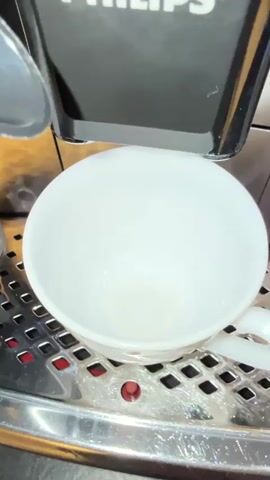 A white ceramic cup sits on a stainless steel grate beneath a black Philips coffee machine. Hot coffee streams from two spouts on the machine into the cup, filling it with a dark liquid. The coffee creates a frothy crema on top as it pours. The background is out of focus, but appears to be a kitchen counter or coffee bar.