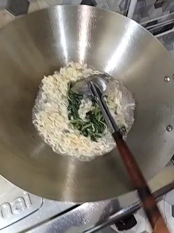 The video shows a close-up shot of a wok on a stovetop. Inside the wok are instant noodles and green leafy vegetables being cooked in bubbling water. A metal spatula with a wooden handle is used to stir the ingredients. The stovetop appears to be a gas stove with a metal grate. The background shows a tiled surface and a light-colored wall. The lighting is bright and direct, creating reflections on the wok. The video is short, lasting only a few seconds, and focuses on the cooking process.