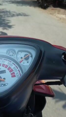 The video begins with a close-up of a red motorcycle's dashboard, the camera angled to show the speedometer and fuel gauge. The perspective suggests the viewer is riding the motorcycle. The scene then shifts to a wider view of a paved road lined with buildings, some appearing to be shops or residences. Sunlight casts shadows from trees onto the road. Several people are visible in the distance, some walking and others near parked motorcycles. One person is seen attending to something near a building with green containers. The video continues to pan across the road, showing more buildings, including one with a gas pump and another with various items displayed outside. The environment appears to be a residential or commercial area in a sunny, outdoor setting.