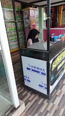 The video shows a street-level view of a food stall. A woman wearing a hijab is standing behind the counter, looking at her phone. The stall has a sign that reads "SOMAY AYAM MALAYA". There are various products and condiments displayed on the counter. In the background, shelves are stocked with what appear to be packaged goods, possibly food items. The ground is tiled. The camera pans to the right, showing more of the street and a parked motorcycle.