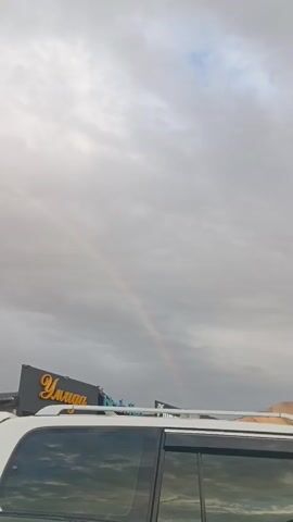 The video shows a landscape with a cloudy sky and a faint rainbow. In the foreground, the side of a white SUV is visible, with reflections of the sky and mountains in its windows. Behind the SUV, a building with a yellow sign is partially visible. Further in the background, a row of cars is parked, and beyond that, mountains are illuminated by sunlight. The overall scene suggests an outdoor setting, possibly a parking lot or roadside area, with a natural phenomenon in the sky.