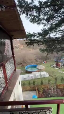 From a balcony, the video shows a backyard scene with a small, round, blue swimming pool surrounded by white lounge chairs and a thatched umbrella. The yard has a green lawn with some artificial turf areas, trees, and a wooden fence. In the background, there's a view of a wooded area. The balcony has a railing and a partially visible structure with a thatched roof. The sky is overcast.