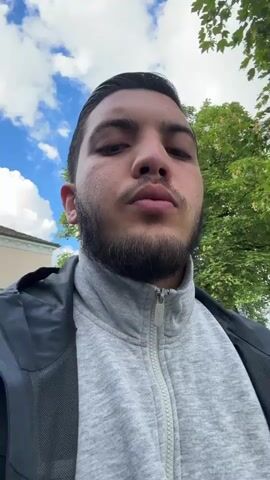 A first-person perspective video shows a young man with dark hair, beard, and olive skin. He is wearing a grey hoodie and a black and blue jacket. The man is outdoors on a sunny day with a partly cloudy sky. Trees and a building are visible in the background. The camera pans slightly, revealing a street with cars and a yellow and white van. The man appears to be talking, but no audio is provided. The video is short, approximately four seconds long.