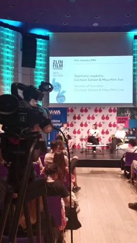 The video shows a live panel discussion at the Zlín Film Festival.  Three individuals are seated on a stage, with an audience seated in front. A large screen displays the event's title: "Film Industry PRO," followed by "Tajemství úspěchu: Cartoon Saloon & Maurfilm Live" and "Secrets of Success: Cartoon Saloon & Maurfilm Live." A professional video camera on a tripod is positioned in the foreground, partially obstructing the view of the stage and audience.  The stage backdrop features a pattern of red teardrop shapes with the number "66" inside.  The room has blue ambient lighting.
