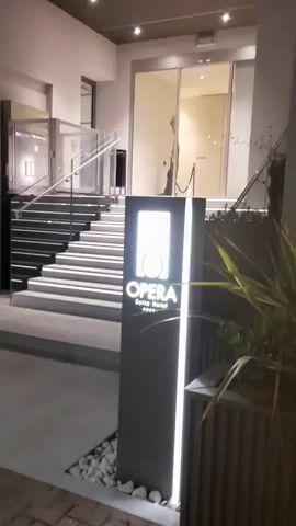 The video shows the exterior of a hotel named "Opera Suites Hotel" at night. The entrance features a staircase with illuminated steps leading to automatic glass doors. A prominent signpost with the hotel's name and stars stands beside the entrance, illuminated by vertical lights. To the right of the entrance, a plant in a planter is visible. The camera then pans to the left, showing a wider view of the street in front of the hotel. Several people are walking on the sidewalk, and there are trees with string lights overhead. In the distance, tables and chairs suggest an outdoor dining area. The sky is dark, indicating it is nighttime.
