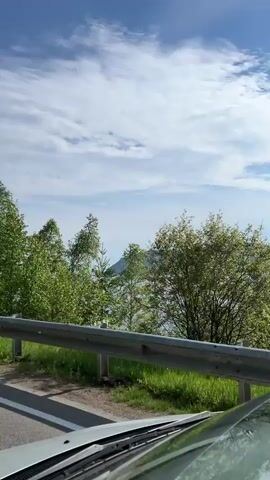 The video shows a view from inside a vehicle moving along a road. The road is paved and has a guardrail on the right side. Beyond the guardrail, there is lush green grass and various trees with dense foliage. The sky is blue with scattered white clouds. The camera appears to be positioned on the dashboard or windshield, and the vehicle is moving forward, causing the trees and landscape to appear blurred due to motion. The scene suggests an outdoor, natural environment, likely during the daytime.