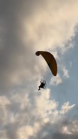 A powered paraglider flies through a cloudy sky. The paraglider, a person attached to a propeller-driven backpack and a wing-shaped parachute, is silhouetted against the sky. The wing is yellow and the clouds are gray and white. The video is filmed from below, looking up at the paraglider. The camera pans slightly, but the paraglider remains the main focus. The overall impression is one of freedom and adventure.
