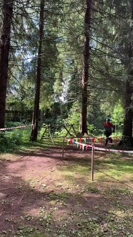 A person wearing a blue helmet and a red and black outfit runs through a wooded area. They are wearing a backpack and a rope is attached to their waist. There are trees and foliage surrounding the person. There are also red and white striped ribbons and flags set up in the area. The person is running from left to right across the screen.