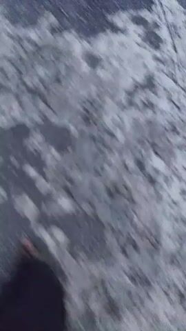 The video shows a first-person perspective of someone walking down a street lined with houses and streetlights. The ground appears to be wet and uneven, possibly due to rain or an unfinished road. The sky is overcast with some visible clouds. The houses are modern in design, with some appearing to be under construction or renovation. The overall atmosphere is calm and quiet, suggesting an early morning or late evening scene.