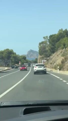 The video shows a view from inside a vehicle driving on a highway. The highway has multiple lanes and is bordered by a steep, rocky slope covered in trees on the right. A light blue sky with no clouds is visible overhead. Several vehicles are on the road, including a white car in the foreground, a red car to the left, and a dark truck further ahead. In the distance, a mountain range is visible against the horizon, and part of the ocean can be seen. Road signs are present, including a speed limit sign showing 50. The overall impression is a scenic drive through a coastal, mountainous region.