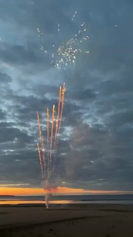 The video shows a fireworks display over a beach and body of water at dusk. The sky is filled with clouds, and the sun is setting, casting an orange glow on the horizon. Fireworks are exploding in the sky, with trails of smoke and sparks descending. The fireworks are predominantly yellow, white, and red. The beach is sandy and appears to be empty of people. There is a body of water in the background, possibly a lake or ocean. The overall mood of the video is festive and celebratory.