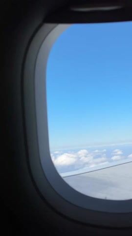 The video shows a view from an airplane window. Outside, a bright blue sky dominates the upper portion of the frame. Below, a layer of fluffy white clouds stretches towards the horizon. Part of the airplane's wing is visible in the bottom right corner of the window frame. The window itself is oval-shaped and framed in gray plastic. The interior surrounding the window is dark.