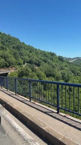 A video captures a roadside view on a bright, sunny day. In the foreground, a blue metal railing borders a concrete structure, casting a shadow on the ground. Beyond the railing, a lush, green hillside is covered with trees. In the distance, a large mountain rises, its peak partially obscured by lighter rock formations. Several small buildings with red roofs are visible scattered amongst the trees on the mountain's lower slopes. The sky above is a clear, vibrant blue. The camera appears to be moving, as indicated by the subtle shifts in perspective.