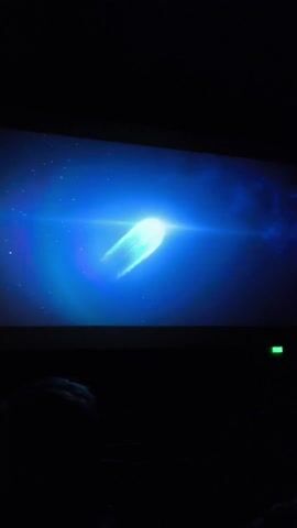 The video was taken in a movie theater, indicated by the silhouette of heads in the dark audience and the exit sign. A bright, animated image is projected on the screen. At first, it looks like a comet. It transforms into what appears to be an abstract spacecraft structure with intricate details, glowing with rainbow colors within a blue and teal aura.