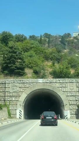 A black sedan drives towards and enters a stone-lined tunnel. The tunnel entrance is arched and decorated with stone carvings. The road is paved and marked with white lines. The surrounding environment appears to be a hilly, vegetated area. The video captures the car's progress through the tunnel, with the interior becoming darker and then brighter as it exits.