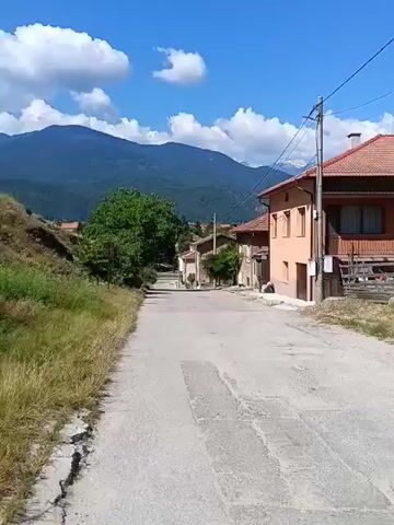 The video shows a view of a village road leading uphill towards a mountain range under a blue sky with white clouds. On the left side of the road, there is a grassy embankment. On the right side, there are houses with terracotta roofs. A large green tree is visible in the middle distance. The road is paved and appears to be in moderate condition. There are no people or vehicles visible in the video. The scene is tranquil and appears to be a rural setting on a sunny day.