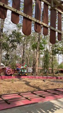 The video shows a toy train with a horse-shaped car on a track in an outdoor park-like setting with trees. The train appears to be moving forward on the track. The front of the train has a red and brown engine with large wheels. The horse-shaped car is green and yellow with a rainbow mane and tail. The track is made of wood and metal and is surrounded by dirt and foliage. The sky is visible in the background, with some clouds. A wooden structure with brown slats is visible in the foreground, obscuring the top of the scene.