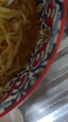 The video shows a close-up, overhead view of a meal. In the foreground, a white rectangular plate holds three pieces of golden-brown French toast. Behind the plate, a colorful bowl with a red, blue, and white patterned rim contains instant noodles in a light-colored broth. A pair of white chopsticks is submerged in the noodles, holding a portion of them. To the left of the bowl, a small portion of a gray mug is visible. The meal appears to be placed on a metallic, ribbed surface, likely a countertop or sink. The lighting is direct and somewhat harsh, casting shadows.