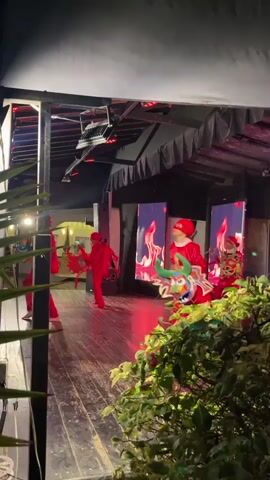 Five individuals in bright red costumes are performing on a wooden stage. The performers are facing forward and to the left of the frame. The performer in the foreground, slightly off-center to the right, is wearing a red costume and holding a colorful, horned mask with green and yellow accents. The other four performers are arranged behind and to the left of this individual, also in red costumes. Some of them appear to be holding props, possibly decorative elements or part of a dance. The stage is covered by a dark, tent-like structure with exposed lighting rigs. Behind the performers, there are large vertical screens displaying fiery, abstract images. Green foliage is visible in the foreground, partially obscuring the view of the stage from the lower left and right corners. The lighting is dim, with the stage area being well-lit by overhead lights and spotlights, casting red hues on the stage and performers. The background shows hints of a building with windows.