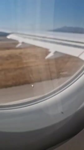 This is a first-person perspective video from inside an airplane window. The view outside the window shows a tarmac with some dry grass and shrubs alongside it. The airplane wing is visible on the right side of the frame. The plane appears to be taxiing or accelerating on the runway. In the distance, there is a control tower and some buildings. The sky is clear and blue.