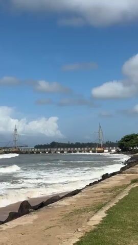 The video shows a scenic view of a coastline on a bright, sunny day with blue skies and scattered white clouds. In the foreground, there is a sandy beach with dark rocks lining the edge of the water. A paved walkway runs parallel to the beach, with green grass beyond it. The sea is visible with moderate waves crashing onto the shore, creating white foam. In the middle ground, there is a long bridge extending across the water. On the left side of the bridge, on a small island covered in lush green trees, stands a white stupa, a Buddhist religious monument. The bridge has a distinctive structure with tall, slender towers on either side, likely supporting the bridge's span. The overall atmosphere is peaceful and picturesque, highlighting a coastal landscape with religious and architectural elements. No people are visible in the video.