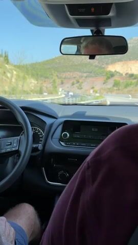 The video shows a view from inside a car as it drives on a road that curves through a hilly, green landscape. The dashboard of the car is visible, including a steering wheel, a radio display showing the time as 15:23, and climate controls. In the rearview mirror, a person's head and shoulders are reflected. The road has guardrails and other vehicles can be seen in the distance. The sky is blue and clear.