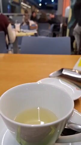 In a close-up shot, clear liquid is poured from a silver teapot into a white teacup filled with a pale-yellow liquid. The cup sits on a white saucer with a spoon placed beside it. The table underneath is wooden. In the out-of-focus background, blurry figures of people are visible sitting at other tables in what appears to be a cafe or restaurant. The teapot has a yellow tag on it, indicating that the tea being poured is chamomile.
