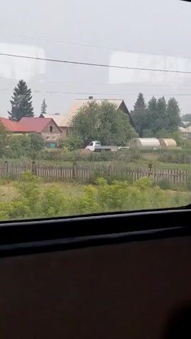 This video shows a view from a window, likely a train or vehicle, looking out at a rural village or countryside. The scene is characterized by houses, some with gardens and fences, and greenery. There are also structures that appear to be greenhouses, wooden fences, and utility poles with wires. The sky is overcast and hazy, suggesting a possibly cloudy or smoky day. The perspective suggests movement, as the scenery changes slightly between frames.