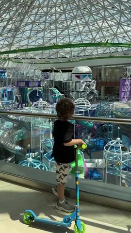 The video captures a young boy with curly brown hair, wearing a black t-shirt and patterned shorts, standing on a blue and green scooter. He is looking out over a large, multi-level indoor playground. The playground is filled with tubes, slides, climbing structures, and ball pits in shades of blue, purple, and white. A large, transparent domed ceiling is visible above, suggesting an indoor space with natural light. The playground appears to be designed with a futuristic or space-themed aesthetic, featuring elements like spherical structures and what looks like a stylized spaceship. The overall impression is one of excitement and wonder as the boy surveys the expansive play area. The location is identified as Sovkhoza imeni Lenina, Russia. The activity is observation of the playground before potential play. The time of day is likely daytime, indicated by the bright natural light filtering through the dome. There is no visible text in the media file.