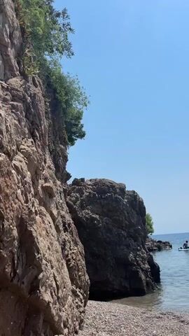 The video shows a rocky coastline with a clear blue sky and calm water. In the foreground, there is a beach covered in pebbles and some driftwood. To the left, a large, textured rock face rises up, partially covered in greenery. In the middle ground, there is another large rock formation emerging from the water. Further out in the sea, a person is visible on a paddleboard. The video pans up the rock face, showing the intricate details of the rock layers and some small plants growing on the surface. The camera then pans back down towards the water and the distant paddleboarder.