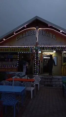 The video shows the exterior of a restaurant called "CHAMBA DHABA PUNJABI RASOI THE BEST FOOD". The restaurant has a prominent sign with lights. Outside the restaurant, there are tables and chairs. A person is walking up the steps into the restaurant. The lighting suggests it is evening or early morning.