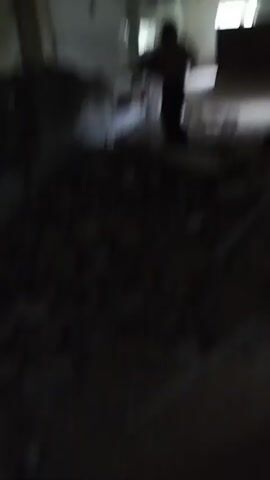 This video depicts a single individual, shirtless and wearing dark pants, engaged in demolition work. They are using a jackhammer to break apart a concrete wall and debris-filled area. The setting appears to be an interior space under renovation or demolition, with rubble and discarded construction materials scattered across the floor. The lighting is dim, suggesting an indoor environment possibly without natural light or with limited artificial illumination. The scene is characterized by dust and the destructive nature of the activity. No text is visible in the video. The time of day and weather are not discernible.