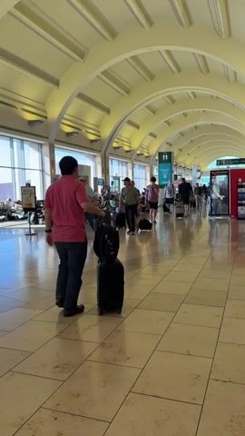 The video shows the interior of an airport terminal. In the foreground, a person wearing a red shirt and blue jeans is walking with a black rolling suitcase and a black backpack. The terminal has a high, arched ceiling and large windows letting in sunlight. People are seen walking in both directions, some with luggage. A Starbucks coffee shop is visible in the middle ground. The floor is tiled with light-colored square tiles. In the background, more people are waiting and walking, and there are signs and displays. The overall atmosphere is that of a busy airport.