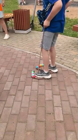 The video shows a person standing on a brick pathway outdoors. The person is wearing a blue t-shirt, grey shorts, and sneakers. They are holding a long stick with strings attached that lead down to several colorful balls arranged in a circle around their feet. The balls are pink, green, yellow, and blue. The person appears to be manipulating the stick, which in turn moves the balls. In the background, there are trees, grass, a wooden bin, and other people walking around. The overall impression is that the person is engaged in some sort of game or activity involving the balls and the stick.