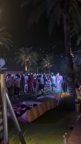 The video shows a large crowd of people gathered at an outdoor party at night. The party takes place in a grassy area with palm trees and string lights overhead. People are standing in groups, some dancing, and many are wearing casual summer attire. In the foreground, there is a striped lounge cushion and various objects on the ground like drinks and metallic poles. The background includes a large white structure and additional trees. The lighting creates a mix of shadows and bright spots, especially on the people and the palm trees.
