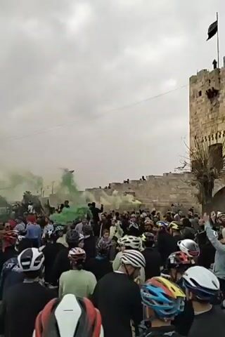 The video depicts a large crowd of people, many wearing cycling helmets, gathered in front of a stone structure that resembles a fortress or ancient wall. Green smoke fills the air, obscuring parts of the scene and adding an element of spectacle or possibly protest. The crowd appears dense, with individuals standing close together and facing in the same direction. Some people are holding bicycles, further suggesting that the event is related to cycling. The sky is overcast, contributing to a somewhat subdued or muted atmosphere. Beyond the crowd and the stone structure, buildings and street lamps can be seen, indicating an urban setting.