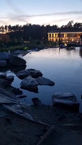 A still shot shows a body of water with rocks scattered around its edge. In the background, a multi-story building with illuminated windows stands behind a line of trees. The sky above is dusky. A waterfall is visible on the right side of the frame, cascading over rocks. Wooden planks are laid out in the foreground.