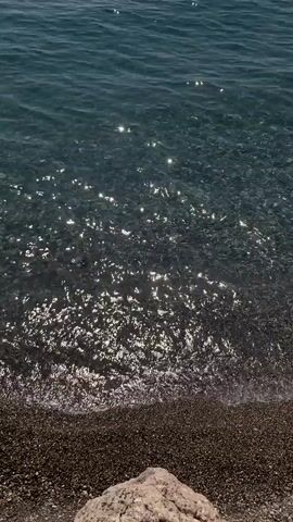 The video is a top-down, stationary shot of a shoreline. The water is clear and blue, with sunlight glinting off the surface. Small waves are gently lapping at the shore, which is covered in dark pebbles and a large, light-colored rock in the foreground. There are no people or other objects visible in the frame.