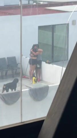 The video captures a daytime scene on a rooftop patio in Cholula, Mexico. A couple, a man and a woman, are embracing each other in a warm hug at the center of the patio. To their left, a black Rottweiler dog slowly walks across the tiled surface. The patio is enclosed by white walls, with a large, dark-framed glass door visible in the background reflection. Several dark outdoor chairs are lined up against one wall, and a red-handled broom leans nearby. Other items include what appears to be a yellow pressure washer and some grey towels draped over the low perimeter wall.