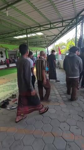 A group of men and boys are gathered outdoors under a covered walkway with a green ceiling. Many of them are wearing traditional Islamic attire, including sarongs and peci hats. Some men are walking in a line, while others are seated on the floor. Several pairs of sandals and shoes are scattered on the ground. In the background, there is a building with green walls and white columns. A bicycle is parked to the right of the frame. The ground is paved with hexagonal tiles. The overall impression is that of a religious gathering or event taking place at a mosque or similar institution.