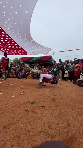 The video shows a group of three men performing a dance in front of a seated crowd. The dancers are wearing matching red and white tops with camouflage pants. The performance takes place on a dirt ground under a large canopy with red and white polka dots. Money is scattered on the ground. The crowd is diverse in age and attire. A few people are standing around the perimeter. The overall atmosphere seems celebratory.
