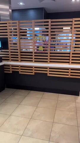 The video shows a close-up view of a McDonald's restaurant interior, focusing on a counter area. A neon sign of french fries with the McDonald's logo is visible behind a wooden slatted screen. The floor is tiled with light brown square tiles. The counter area has a dark base with a white surface.