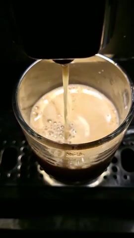 A close-up shot shows a stream of milk being poured into a glass mug filled with coffee. The milk creates a swirling pattern as it mixes with the coffee, forming a light brown frothy mixture. The mug sits on a perforated metal drip tray, suggesting it is part of an espresso machine. The lighting is dim, with the focus on the coffee and milk interaction.