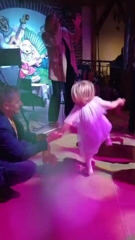 The video shows an indoor scene with a dance floor and a stage. A woman is singing on the stage, and a young girl in a white dress is dancing in front of her. There are other people in the background, some of whom are also dancing. The lighting is dim and colorful, with purple and green lights illuminating the scene. A mural depicting cartoon characters playing instruments is visible on the back wall of the stage. A music stand is placed in front of the stage. A person is sitting on the floor in the foreground, facing the girl.