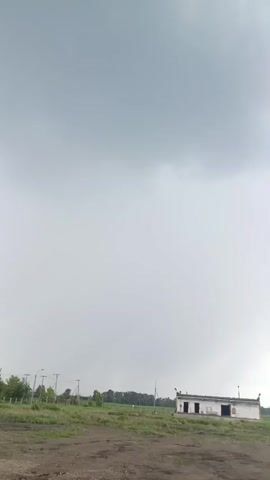 This is a vertical video of an outdoor scene under a cloudy sky. The foreground shows a dirt path and some patchy grass. In the middle ground, there are several structures. On the right, a small, white building with two dark doorways is visible. To the left of it, a grassy area is bordered by a fence with utility poles and lights. Further to the left, another small, white building or shed is partially visible, with some wooden pallets stacked nearby. The background consists of a line of trees and a vast expanse of grey clouds. The weather appears overcast, suggesting it might be a gloomy or rainy day. There is no visible text in the video. The overall mood is subdued, possibly reflecting the weather conditions.