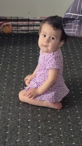 A baby wearing a purple polka dot dress is sitting on a dark patterned carpet. The baby turns their head to the right as if looking at something off-camera.