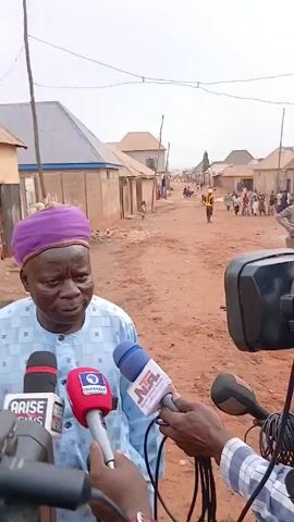 A man is being interviewed by multiple media outlets. He is wearing a light blue patterned shirt and a purple head wrap. Four microphones are visible, each from different news organizations: Arise News, Channels, and NTA. The interview is taking place outdoors on a dirt road, in a village-like setting with small, simple buildings in the background. There are a few people in the distance and in the mid ground a small child. The man is looking at the interviewers. The interviewers are holding their microphones towards the man.