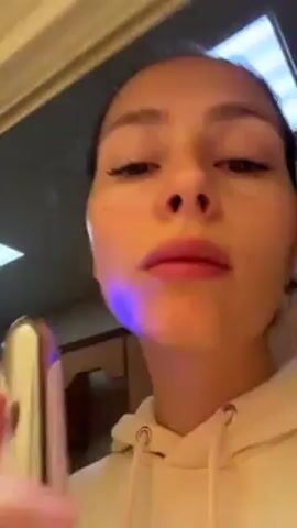 Here is a detailed description of the video:
The video shows a woman holding a device in her right hand. The device is slim, with a light pink body and a gold-colored metal applicator end that emits a blue light. The woman is using the device on her face, moving it along her cheek, under her eye, and along her nose. She looks directly at the camera, focusing on her face in a mirror. She is wearing a cream-colored hoodie. The background shows a bathroom interior with light-colored walls, a light fixture in the ceiling, and a glimpse of a wooden cabinet.
