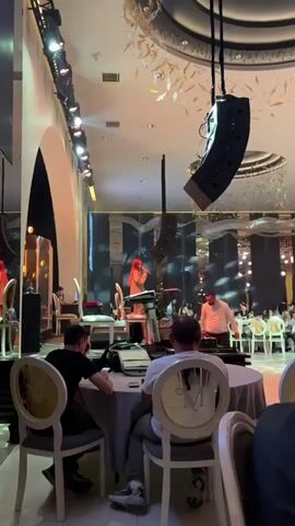 A video captures a live musical performance within a large, ornate hall, likely a wedding venue. A woman with red hair, wearing a beige outfit, sings into a microphone while standing behind a Roland keyboard on a raised stage. The background features dark, mirrored panels accented with warm lighting. Suspended from the ceiling above the stage is a large, black, box-shaped speaker. Below the stage, attendees are seated at round tables, some raising their hands in enjoyment. A man is visible in the foreground working at an audio mixing board at a table. The lighting in the hall is warm and decorative, and the overall atmosphere suggests an upscale event.